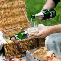 Ein Mann gießt Sekt in ein Glas, während er an einem Picknickkorb auf einer grünen Wiese sitzt.