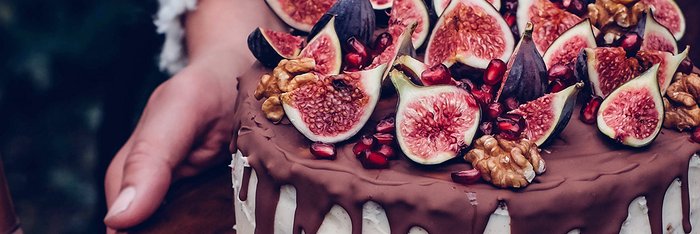 Der Kuchen, überzogen mit Schokolade, wird von einer Hand gehalten, während frische Feigen und Nüsse darauf arrangiert sind.