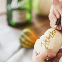 Eine Hand schreibt mit einem goldenen Stift das Wort 'Herbst' auf einen weißen Stein, umgeben von herbstlichen Dekorationen.