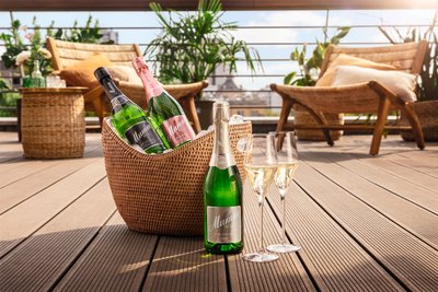 Auf einem Rillen-Boden steht eine Sektflasche und zwei Sektglser sowie dahinter ein Sektkühler, im hinteren Hintergrund ist eine Sommerterrasse zu antizipieren