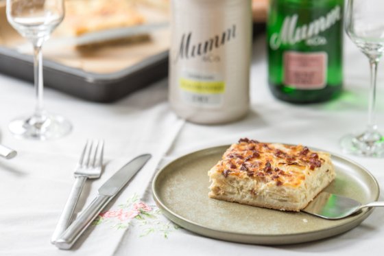 Ein Stück herzhaftes Zwiebelkuchen liegt auf einem Teller, umgeben von Gläsern und Flaschen auf einem elegant gedeckten Tisch.