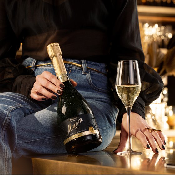 Die Hand einer Person mit schwarzem Nagellack umschließt eine Flasche Mumm, während ein elegantes Glas mit sprudelndem Sekt bereitsteht.