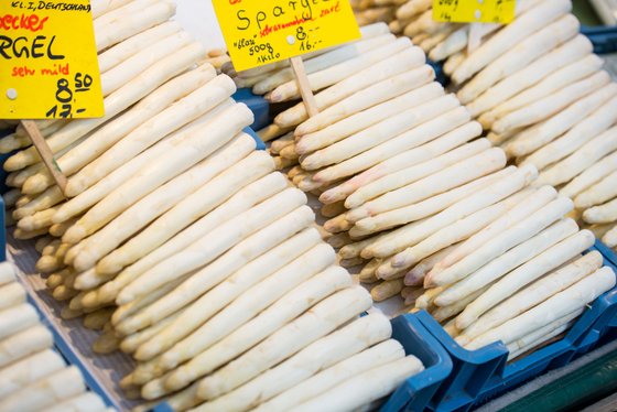 Frischer, weißer Spargel liegt ordentlich in blauen Kisten, umgeben von bunten Preisschildern auf einem Marktstand.