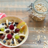 In einer hübschen Schüssel vereinen sich Joghurt, Bananen, Trauben und Himbeeren, während ein Glas Haferflocken daneben steht.