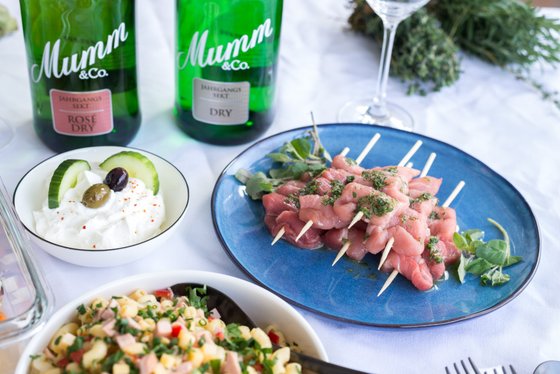 Zwei Flaschen Mumm Sekt stehen im Hintergrund, während im Vordergrund ein Teller mit saftigen Fleischspießen und frischen Kräutern zu sehen ist.