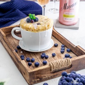 Eine weiße Tasse mit einem überlaufenden Dessert, garniert mit frischen Blaubeeren und einem Minzblatt, auf einem Holztablett.