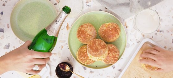 Auf dem Tisch liegen goldbraune scones, während eine Hand eine Sektflasche öffnet, um einen festlichen Moment zu feiern.