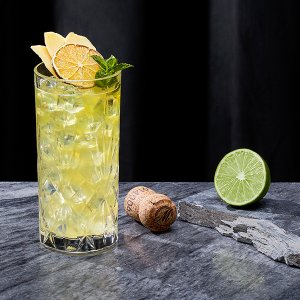 Ein gläserner Cocktail mit Eiswürfeln, dekoriert mit Zitronen und Minze, neben einer halbierten Limette und einem Korken.