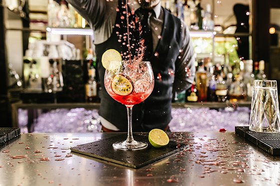 Ein leuchtend roter Cocktail mit Limetten wird von einem Barkeeper in einem schicken Anzug zubereitet, umgeben von Flaschen.