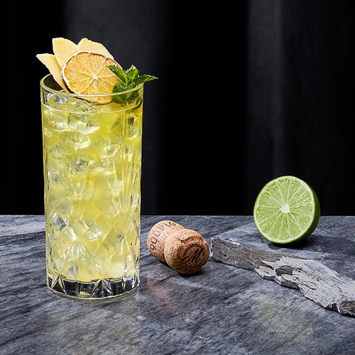 Ein gläserner Cocktail mit Eiswürfeln, dekoriert mit Zitronen und Minze, neben einer halbierten Limette und einem Korken.