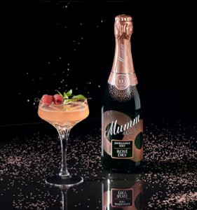 Ein elegantes Glas mit roséfarbenem Cocktail, garniert mit Himbeeren und Minze, steht neben einer Flasche Mumm Sekt.