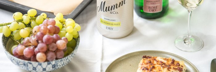 Ein elegantes Tischarrangement mit einer Schüssel Trauben, alkoholfreiem Sekt und einem köstlichen Stück Zwiebelkuchen auf einem Teller.