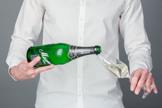 Eine Person in einem weißen Hemd gießt Sekt aus einer grünen Flasche in ein elegantes Glas.