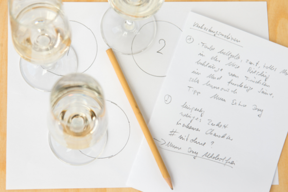 Drei Weingläser stehen auf einem Tisch, umgeben von Notizen und einem Bleistift, der auf einem Blatt Papier liegt.