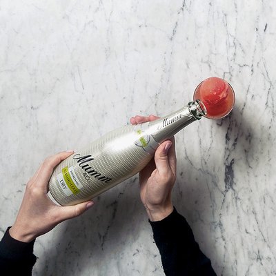 Die Hände einer Person umschließen eine silberne Sektflasche, während ein Glas mit rotem Sorbet darauf platziert ist.
