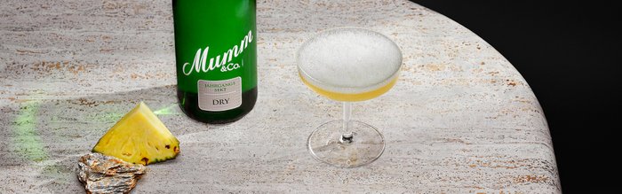 Auf einem hellen Tisch liegt eine Flasche Mumm Sekt, daneben ein Cocktailglas mit schaumigem Getränk und einer Ananasscheibe.