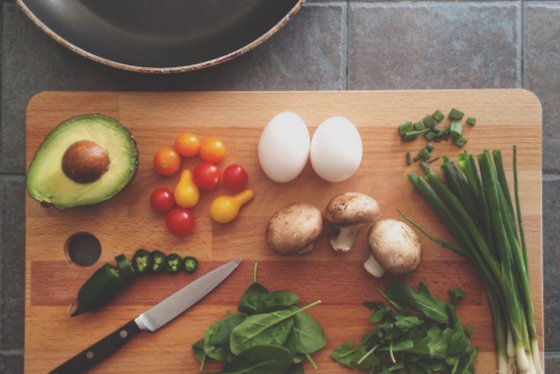 Auf einem Holzbrett liegen frische Zutaten: Avocado, Eier, Tomaten, Pilze und Gemüse, bereit zum Kochen.