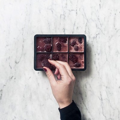Auf einem Marmoruntergrund liegt eine Eiswürfelform mit gefrorenen Brombeeren, während eine Hand eine Frucht herausnimmt.