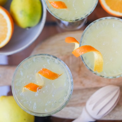 Auf einem Holzbrett stehen drei Gläser mit einem gelben Cocktail, dekoriert mit Orangenschalen und umgeben von Zitronen und Orangen.