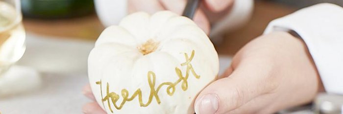 Eine Person hält einen weißen Kürbis, auf dem mit goldener Farbe das Wort 'Herbst' geschrieben steht.