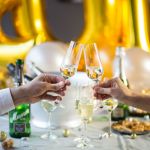 Zwei Hände stoßen mit Sektgläsern an, umgeben von festlicher Dekoration und goldenen Luftballons.