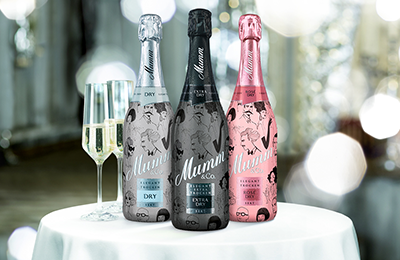 Auf einem Tisch stehen drei Flaschen Mumm Sekt, eine in Schwarz, eine in Silber und eine in Rosa, neben einem Glas Sekt.