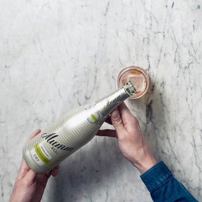Eine Person hält eine Flasche Mumm-Sekt über einem Glas mit einem sprudelnden Getränk auf einer marmorierten Oberfläche.