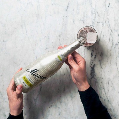 Eine Person hält eine Flasche Mumm Sekt mit einem Glas, das einen Eiswürfel enthält, vor einer marmorierten Oberfläche.