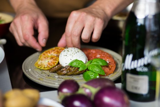 Eine Hand platziert frische Tomatenscheiben und eine cremige Burrata auf einem Teller, garniert mit Basilikumblättern.