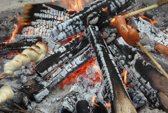 Glühende Holzscheite und glühende Kohlen bilden ein lebhaftes Lagerfeuer, während Würstchen und Teigspieße darauf grillen.