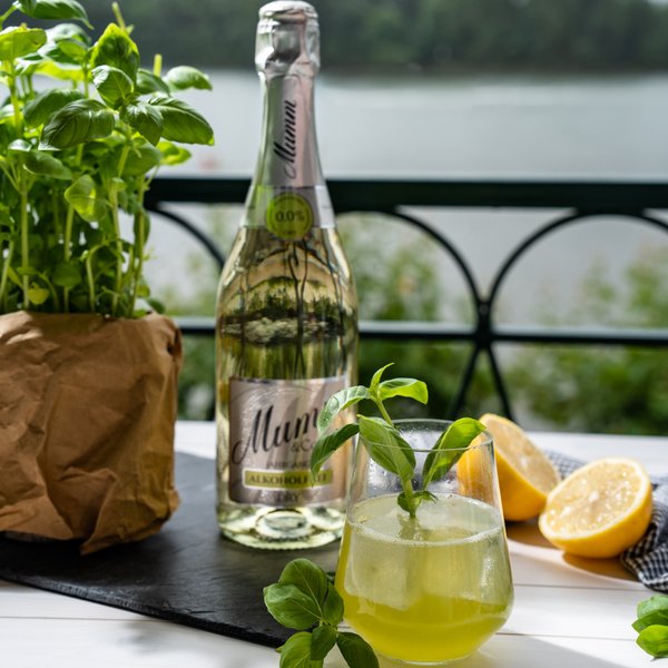 Ein sonniger Tisch mit einem Glas Getränk, einer Flasche Mumm und frischen Zitronen, umgeben von üppigem Grün am Wasser.