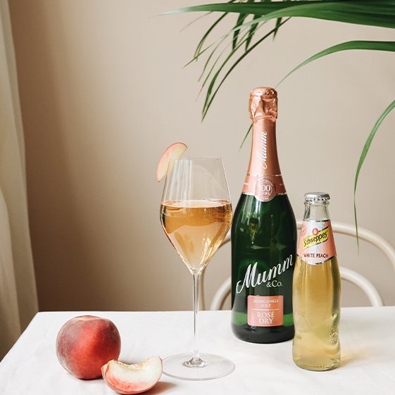 Ein stilvolles Arrangement mit einem Glas Rosé-Sekt, einer Flasche Mumm und einem frischen Pfirsich auf einem weißen Tisch.