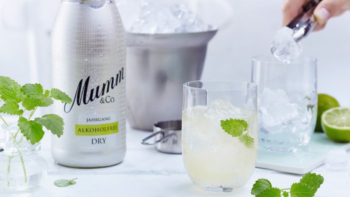 Ein Glas mit sprudelndem Getränk, gefüllt mit Eis und frischer Minze, während eine Hand Eis hinzufügt.