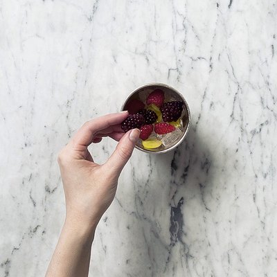 Eine Hand greift nach frischen Beeren in einer silbernen Schüssel, die auf einer marmorierten Oberfläche steht.