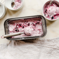 Ein offenes Eisbehälter mit lila Beeren-Eis, daneben zwei leere Schalen und Löffel auf einem hellen Tisch.