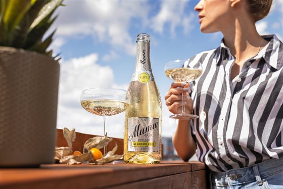 Eine Person in grau-weiß gestreiftem Oberteil steht an einem Tisch mit zwei gefüllten Gläsern und einer Flasche Sekt sowie einer Pflanze
