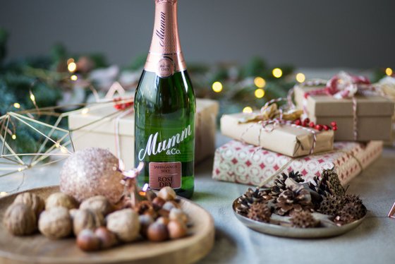 Eine Flasche Mumm Sekt steht vor einer festlich dekorierten Kulisse mit Geschenken und Weihnachtslichtern.