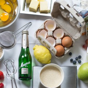 Eine Vielzahl von Zutaten liegt auf einem Tisch, darunter frische Eier, Obst und eine Flasche Sekt, bereit zum Kochen.