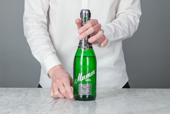 Der Fokus liegt auf einer Hand, die eine Flasche Mumm Extra Dry hält, während die andere Hand den Korken greift.