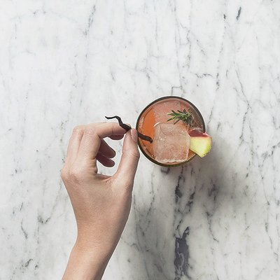 Eine Hand hält ein Glas mit einem bunten Cocktail, das mit einem Eiswürfel und frischen Zutaten verziert ist.