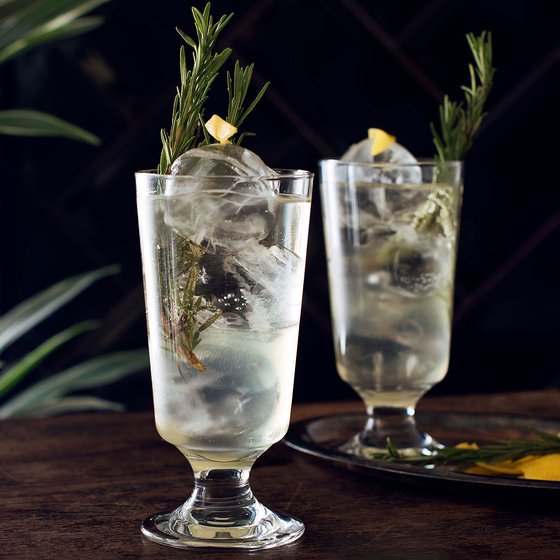 Zwei elegante Gläser mit einem sprudelnden Getränk, gefüllt mit Eis und frischen Rosmarinzweigen, auf einem Holztisch.