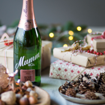 Eine Flasche Mumm Sekt steht vor festlich verpackten Geschenken und einer dekorativen Tischlandschaft mit Tannenzapfen.