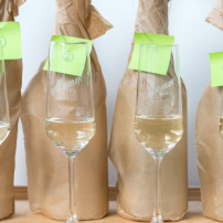 Vier Flaschen Sekt in braunem Papier verpackt, stehen neben eleganten Gläsern auf einem Holztablett.
