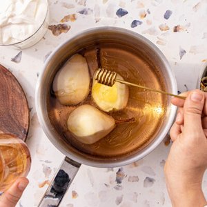 In einem Topf werden drei Birnen in einer goldenen Flüssigkeit gekocht, während ein Löffel Honig darüber geträufelt wird.