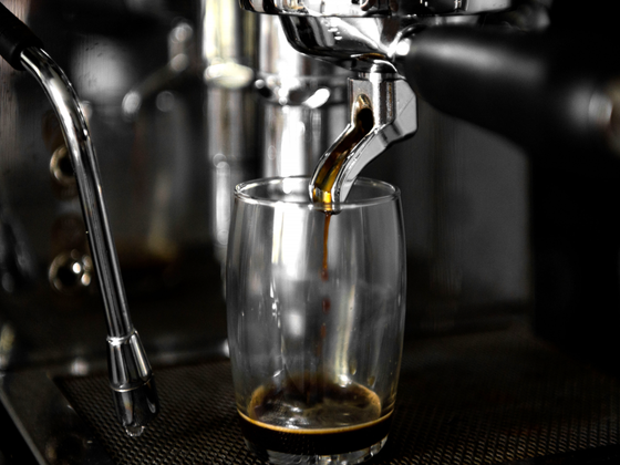 Der Kaffee fließt aus der Maschine in ein Glas, das auf einer dunklen, strukturierten Oberfläche steht, umgeben von glänzendem Metall.