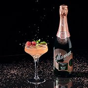 Ein elegantes Cocktailglas mit einem rosafarbenen Getränk, garniert mit Himbeeren und Minze, steht neben einer Flasche Sekt.