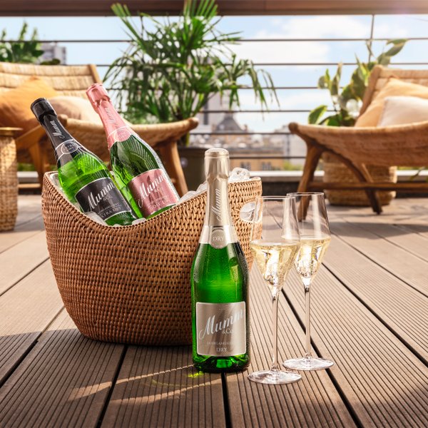 Auf einem Rillen-Boden steht eine Sektflasche und zwei Sektglser sowie dahinter ein Sektkühler, im hinteren Hintergrund ist eine Sommerterrasse zu antizipieren