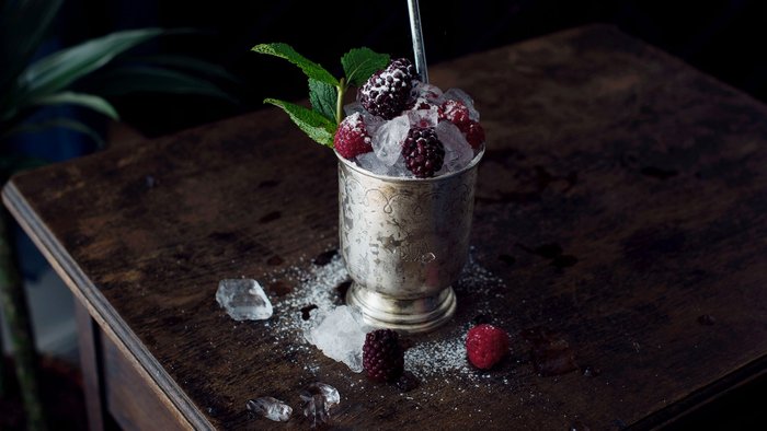 Ein silberner Becher gefüllt mit Eis, Himbeeren und Brombeeren, steht auf einem dunklen Holztisch, umgeben von Eisstücken.