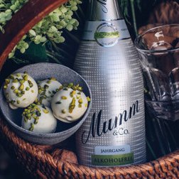 In einem geflochtenen Korb liegen eine Flasche Mumm, ein Glas und zarte Pralinen, die mit grünen Pistazien bestreut sind.
