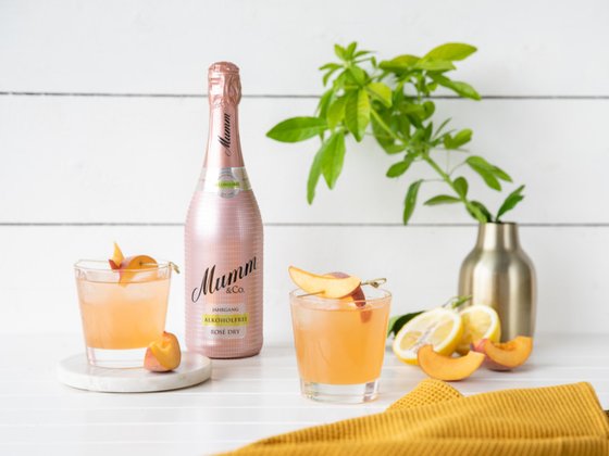 Zwei Gläser mit einem erfrischenden, roséfarbenen Getränk, garniert mit Pfirsichscheiben, stehen neben einer Flasche Mumm.